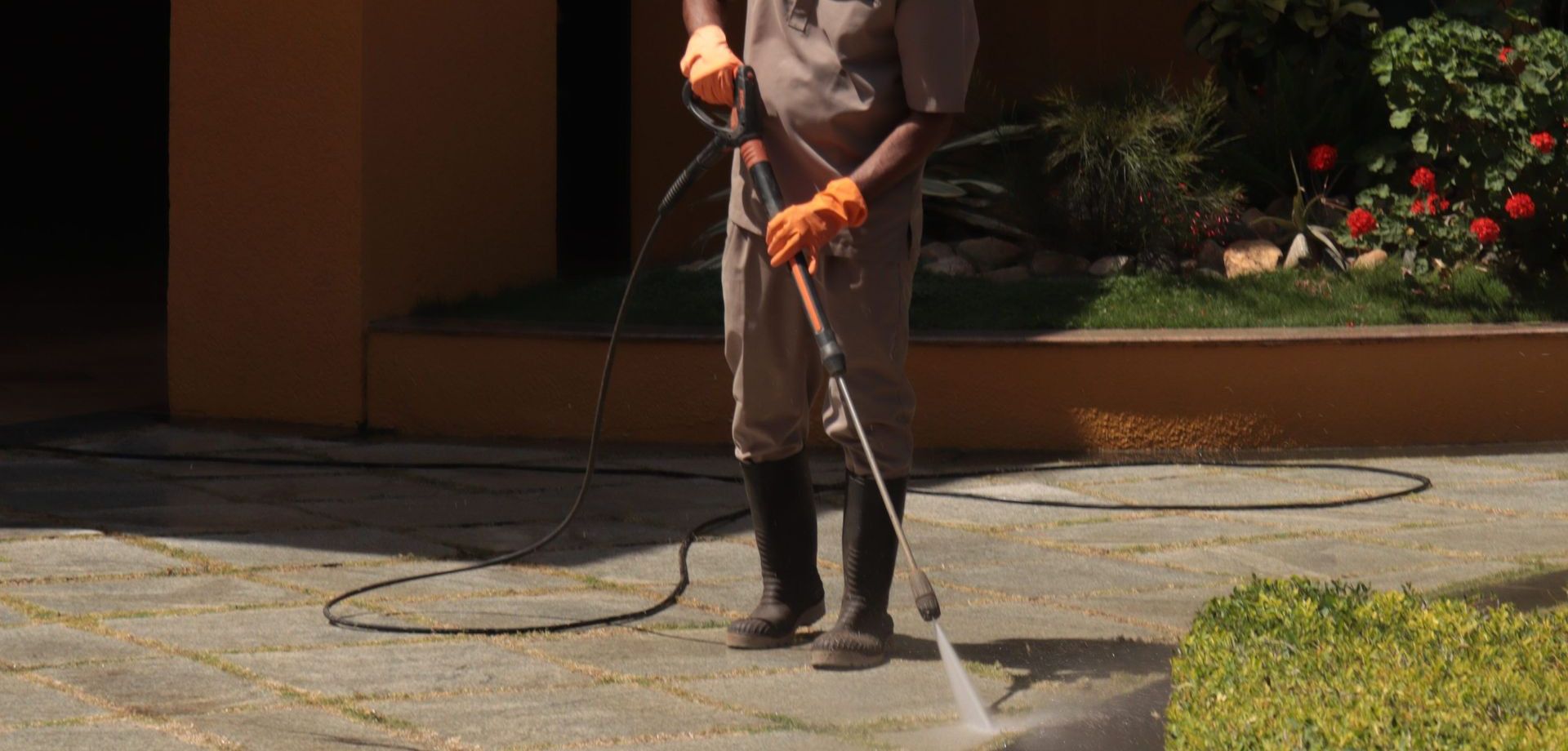 A gardener wearing a hat cleans an urban sidewalk with a pressure washer.